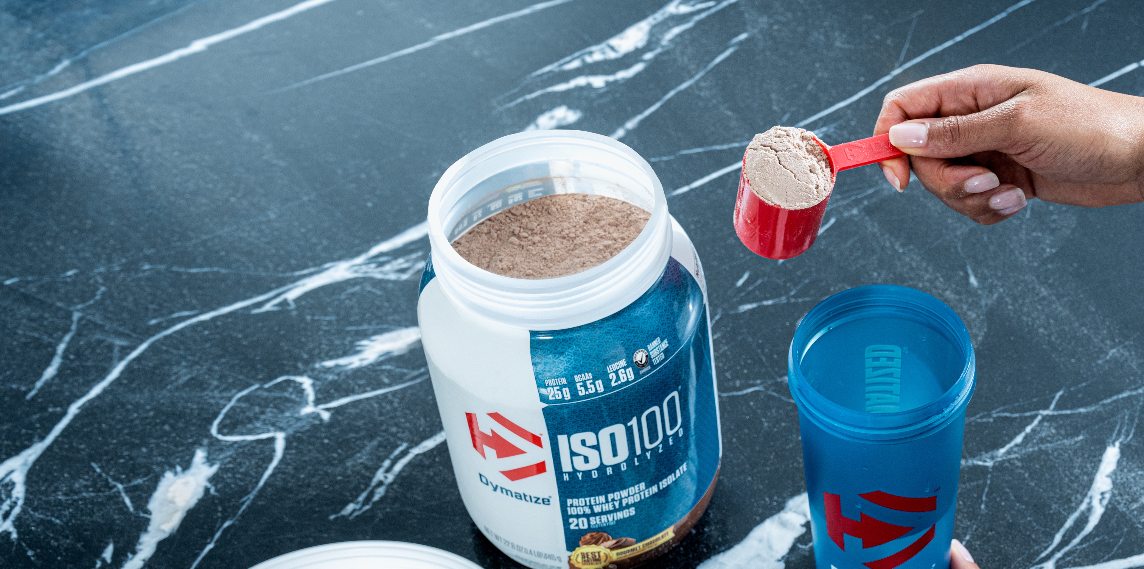 A single tub of Dymatize ISO100 open, with a hand pulling a scoop of powder out in a scooper.
